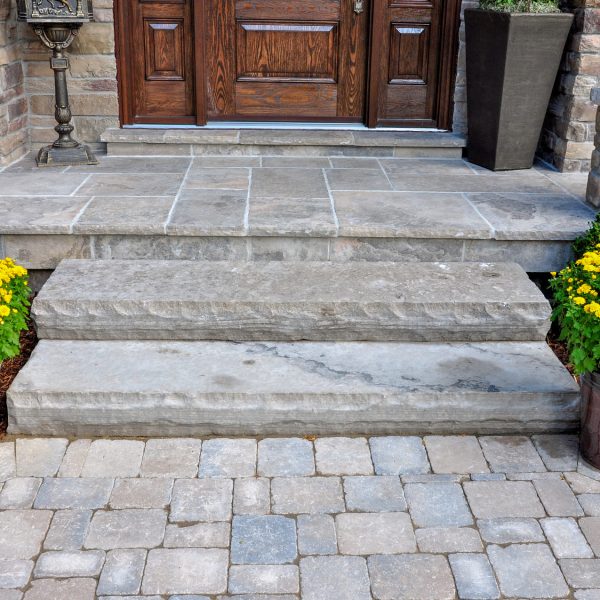 Stone steps lead to a wooden front door, flanked by yellow flowers in pots and brick pavers